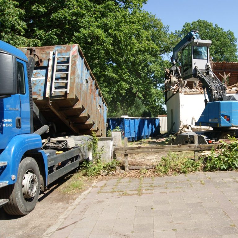 Zwei Müllfahrzeuge stehen in einem befestigten Bereich umgeben von Bäumen.